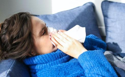 A woman with a flu is lying in the couch blowing her nose.