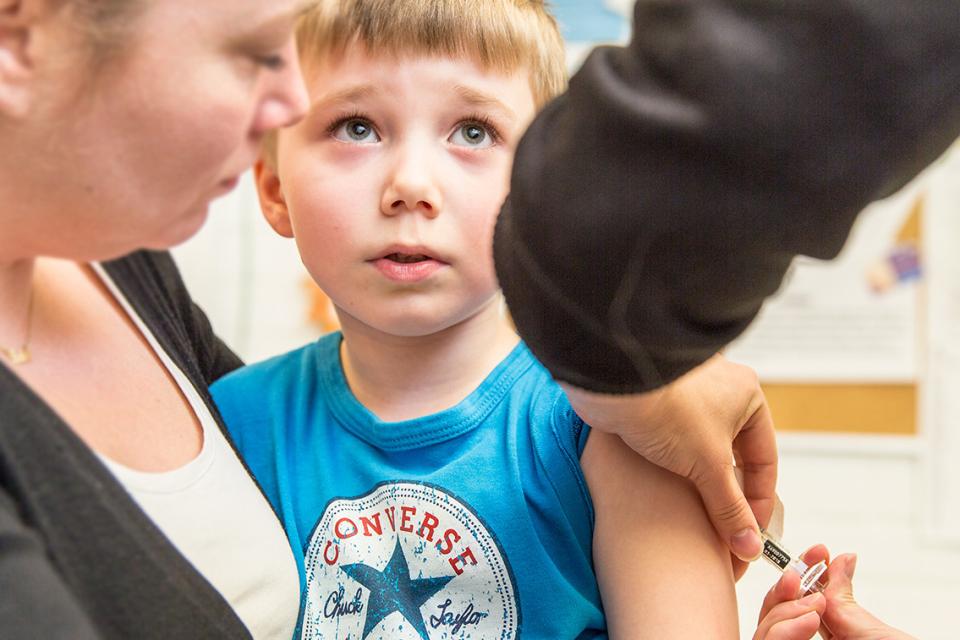 Barnet sitter i en förälders knä. Framför barnet ses händerna som lägger vaccinet i barnets arm. Barnets blick riktas mot vaccinatorn.