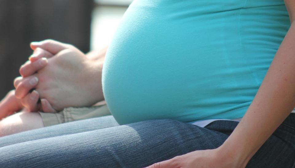 Magen på en gravid kvinna. Kvinnan är klädd i en turkos skjorta och jeans. Hon sitter på en bänk och håller en annan persons hand.