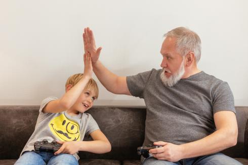 Nuori poika ja vanha, harmaantunut mies heittävät läpyt sohvalla videopeliohjaimet syleissään.