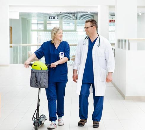 Two people are walking in the hospital's white corridor. They are wearing hospital work clothes. They look at each other and one pushes the kickboard.