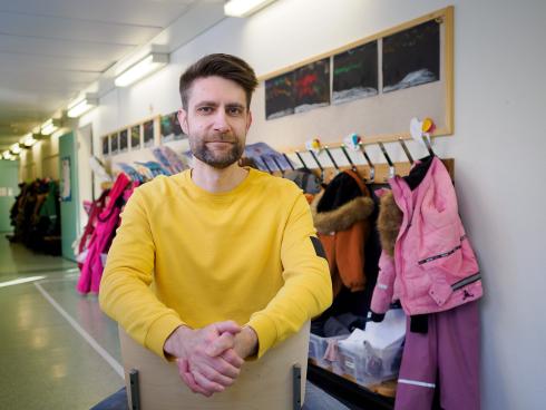 A person sits in the school corridor and leans on the back of a chair facing the wrong way. The person is wearing a yellow shirt and is looking at the camera. In the background are children's clothes on a coat rack.