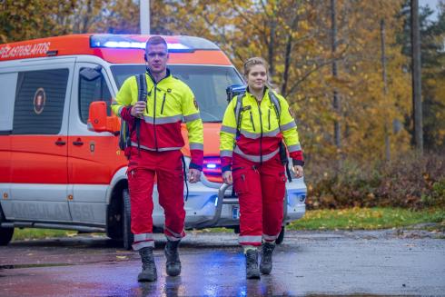 A fireman and a paramedic walking towards the camera. An ambulance is parked behind them.