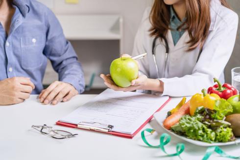 Terveysalan ammattilainen valkoisessa takissa istuu pöydän ääressä asiakkaan kanssa. Hän pitää kädessään omenaa. Pöydällä on papereita, lautasella vihanneksia ja mittanauha.