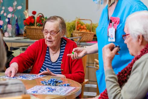 Kaksi iäkästä naista pelaa bingoa ja hoitaja seisoo taustalla.