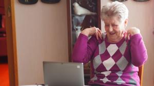 Ikääntynyt nainen istuu kannettavan tietokoneen edessä ja tekee hartiajumppaa.