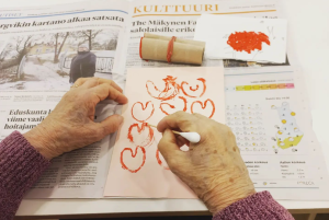 Vanhan ihmisen käsissä on kynä, jolla hän piirtää punaisia sydämiä valkoiselle paperille. Paperin alle on levitelty avoin sanomalehti. 
