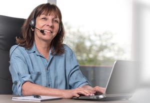 En glad kvinna sitter vid datorn med hörlurar på huvudet och svarar i telefonen..