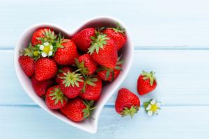 Jordgubbar i en hjärtformad behållare. Bilden tagen från ovan. Bredvid fatet, på ett ljust träbord, två jordgubbar och en jordgubbsblomma.