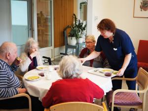 Tummansinisiin vaatteisiin pukeutunut hoitaja tarjoilee lounasta neljän hengen pöytäseurueelle ikääntyneiden palveluasumisen yksikössä.