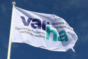 Valkoinen lippu lipputangossa. Lipussa on logo, jossa lukee ”Varha. Varsinais-Suomen hyvinvointialue. Egentliga-Finlands välfärdsområde”. Taivas taustalla on kirkkaan sininen.