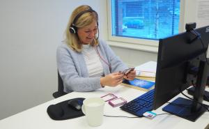 En blond kvinna sitter vid datorn med hörlurar i öronen.