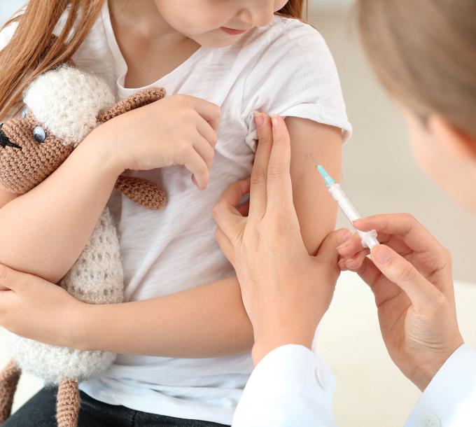 Läkare lägger ett vaccin i barns arm. Barnet har en nalle i famnen.