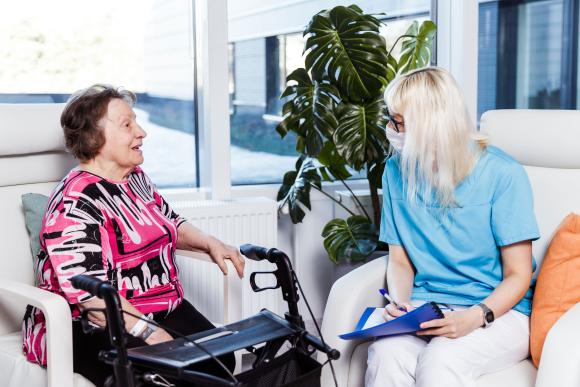 Iäkäs nainen juttelee hoitajan kanssa. Kumpikin istuu tuolissa ja iäkkäällä naisella on edessään rollaattori.