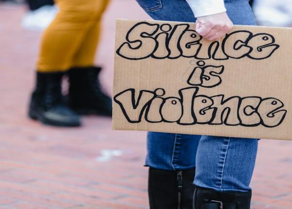 Henkilö, jolla on kädessä pahvikyltti, jossa lukee "Silence is violence".