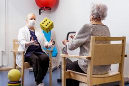 En läkare och en äldre person sitter på stolar med svar. De slår en stor tärning mot varandra.