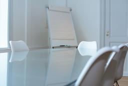 Lasinen suorakaiteen muotoinen pöytä, jonka ympärillä valkoisia tuoleja. Taustalla huoneen nurkassa fläppitaulu.