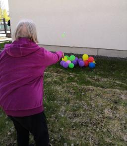 Harmaahiuksinen nainen yrittää osua heittämällään tikalla maassa oleviin ilmapalloihin