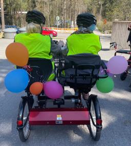 Kahdenistuttavan pyörän ohjaimissa kaksi henkilöä kuvattuna selkä päin. Heillä on ilmapalloja koristeenaan. 