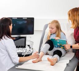 Ett barn sitter på en sjukhussäng. En person med vit jacka undersöker hennes ben och har en dator framför sig. Bredvid barnet finns en vuxen.