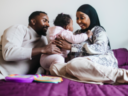 Muslimiperheen iloiset isä, äiti ja vauva leikkivät sängyllä ja hymyilevät.