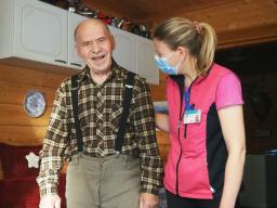 En gammal man i en rutig skjorta står i sitt hem och skrattar. Bredvid honom står en kvinnlig närvårdare med munskydd.