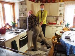 En gammal man lutar sig mot en köksbänk i sitt hem och utför sparkrörelser bakåt. Bredvid honom står en manlig fysioterapeut och leder verksamheten.